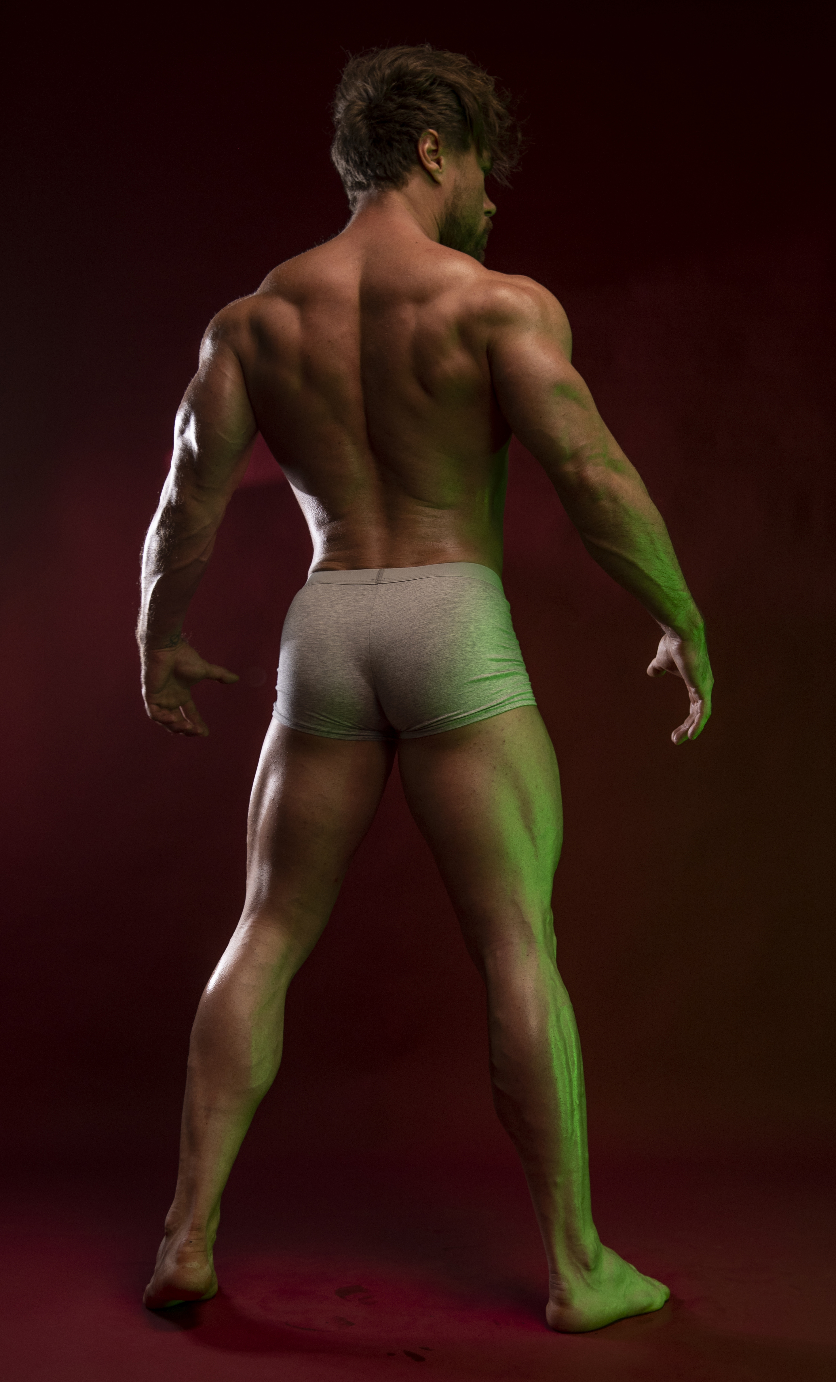 a muscular man with a defined back and legs stands in underwear under green lighting in a studio setting.