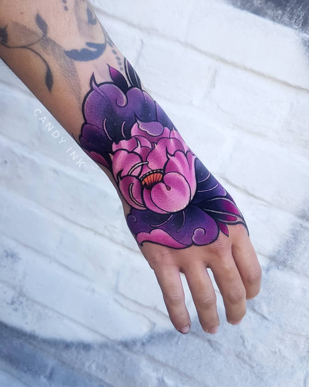 a closeup photograph showcasing a freshly inked tattoo on an individual's hand. the tattoo, rendered in a vibrant palette of pinks and purples, depicts a stylized flower, likely a peony, with accompanying leaves and cloudlike accents. the art style leans towards neotraditional, with bold lines and smooth shading. the setting suggests a tattoo studio, evident from the clean, white brick wall backdrop and the soft, diffused lighting that accentuates the tattoo's details.