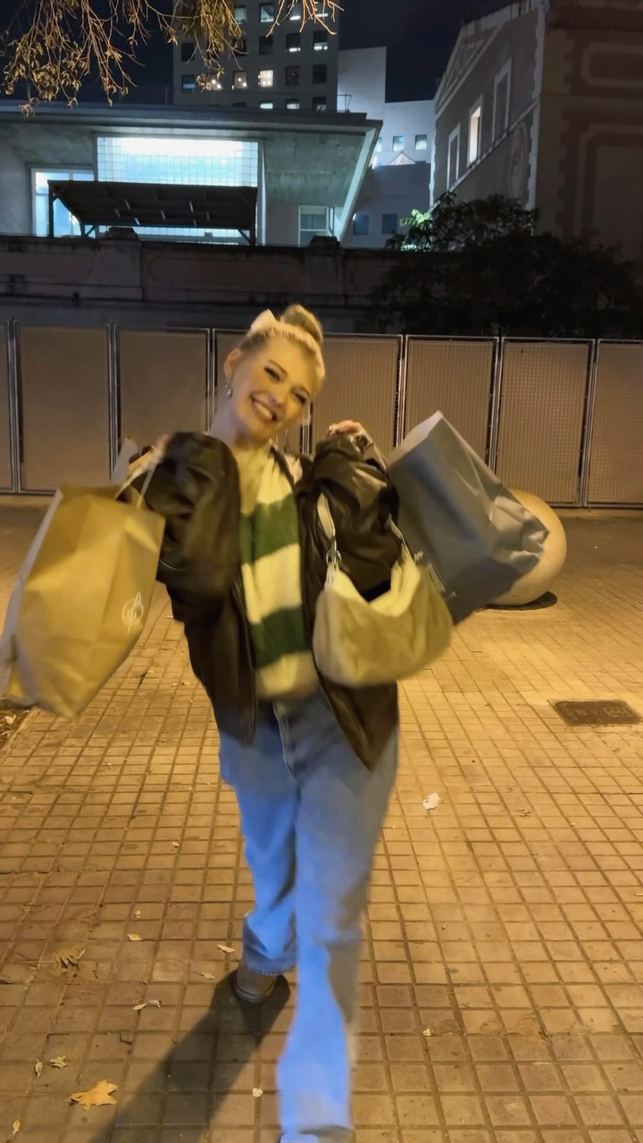 a smiling woman stands on a tiled sidewalk at night, holding multiple shopping bags and a purse. she is wearing jeans and a jacket over a striped sweater. buildings are visible in the background under a dark sky.