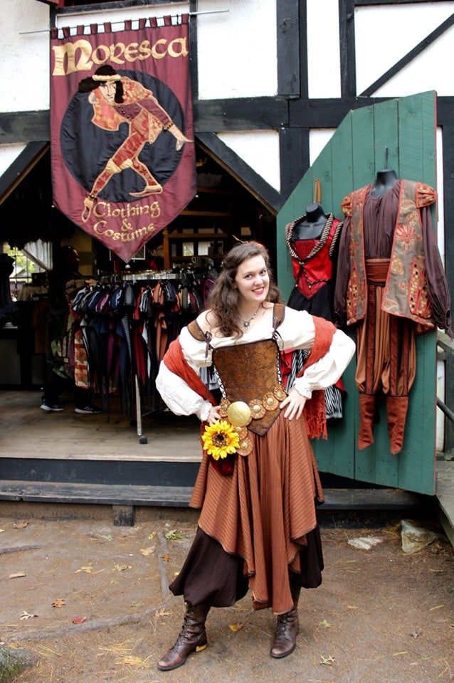 a woman dressed in a medieval costume stands in front of a clothing store at a renaissance fair. she is wearing a brown corset, a white blouse, a brown skirt, and brown boots. she has a sunflower pinned to her belt. behind her is a store selling medieval clothing and costumes. a banner for the store hangs above the entrance. there are clothing racks inside and display outfits hanging by the door.