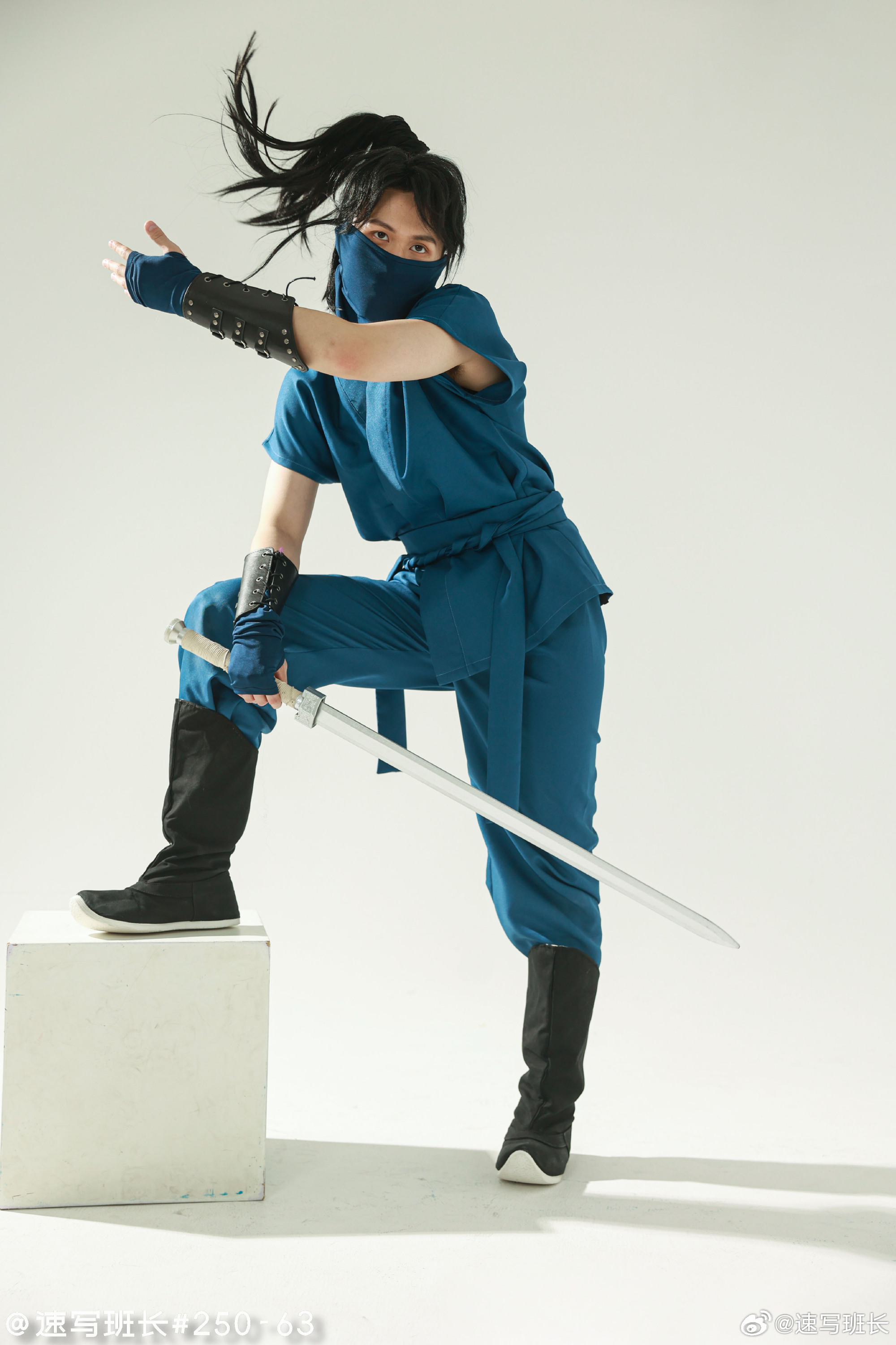 a person dressed as a ninja in a blue outfit and a black face mask is striking a pose on a white background, wielding a sword. their long black hair is blowing in the wind, created by a fan off camera.