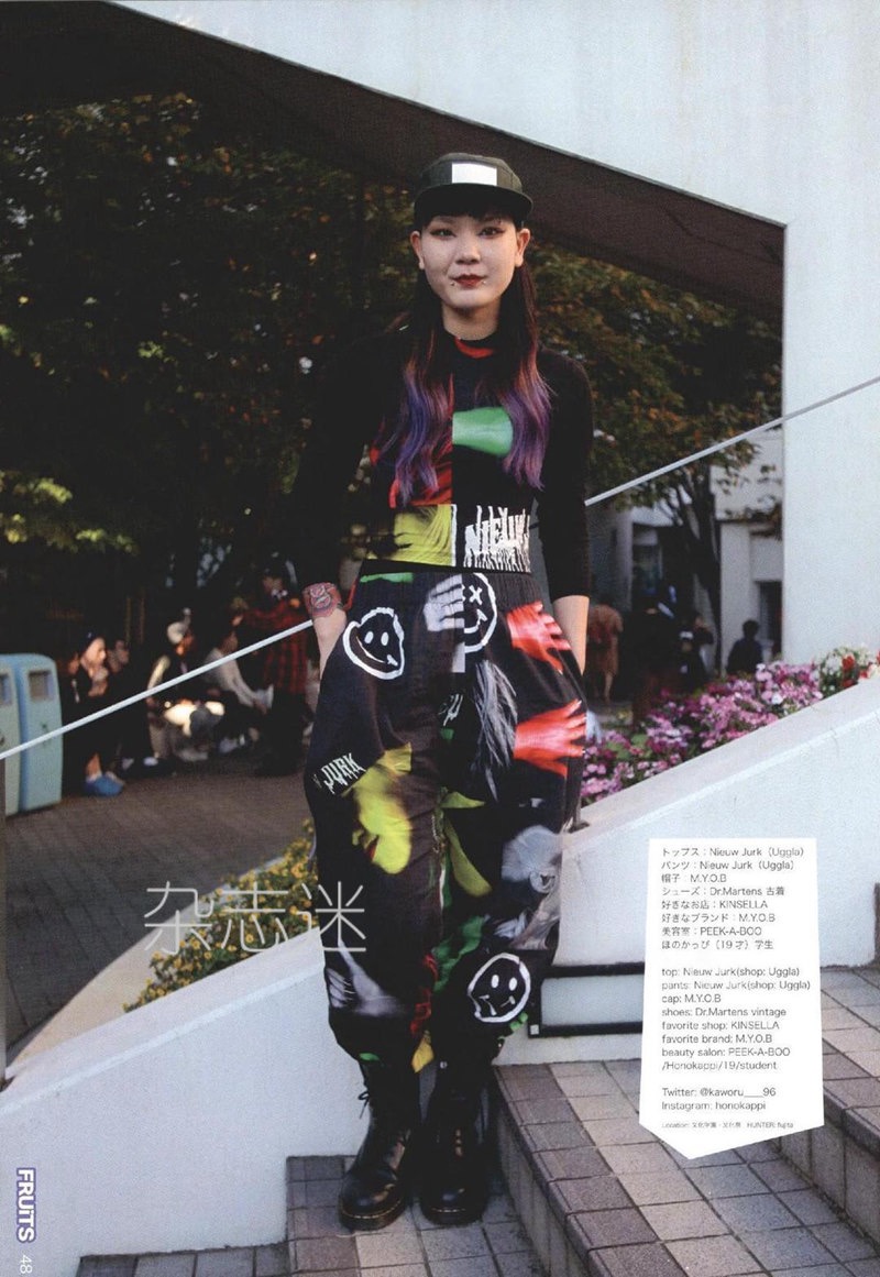 a young woman with purple hair is posing on a set of stairs in an urban environment. she is wearing a black longsleeved shirt, colorful printed overalls featuring smiley faces and hands, and black boots. her outfit showcases a streetwear style popular in harajuku, tokyo. the background features buildings, greenery, and other people walking by.