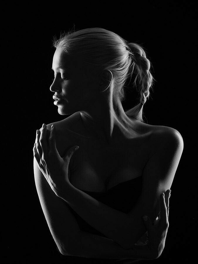 a woman in a black and white portrait is illuminated from the right side of her face and shoulders, her eyes and lips are visible. she is standing in a dark background and is wearing a simple black top. her hair is pulled back in a braid, and she is looking down and to her left. she appears to be lost in thought and her hands are clasped together on her chest. the image has a mysterious and sensual feel.