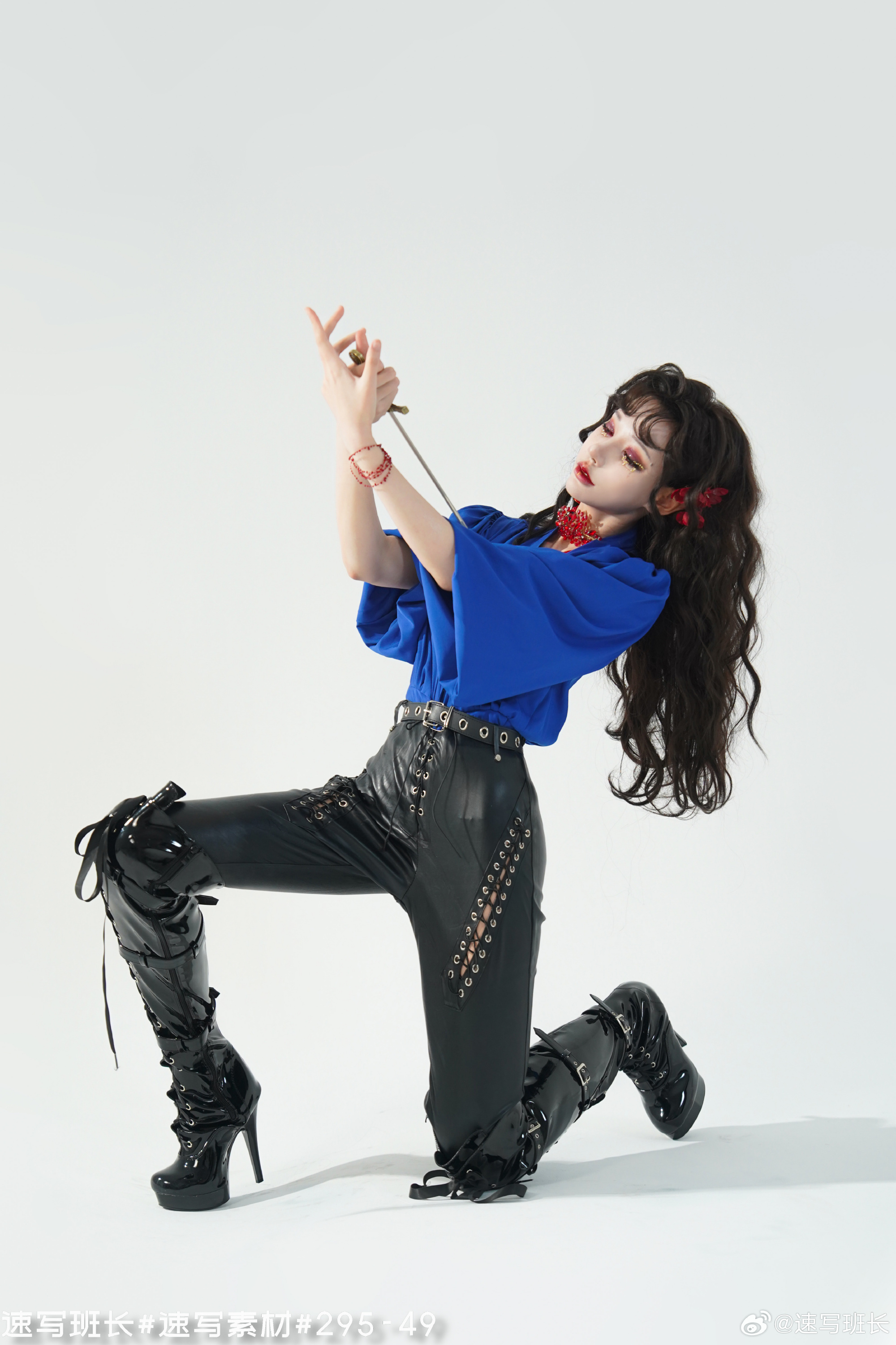 a woman with long black hair wearing a blue shirt, black leather pants, and black highheeled boots is posing in a studio. she is holding a sword in her right hand and has her left hand raised above her head. she is looking up at the sword. the background is white.