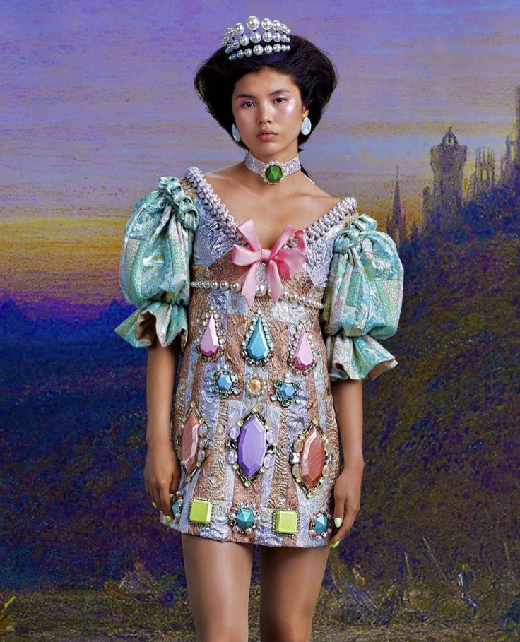 a young woman wearing a princess costume. she's wearing a short, ornate dress decorated with colorful jewels. she's also wearing a jeweled crown and earrings. the background is a colorful painting with castle silhouettes.