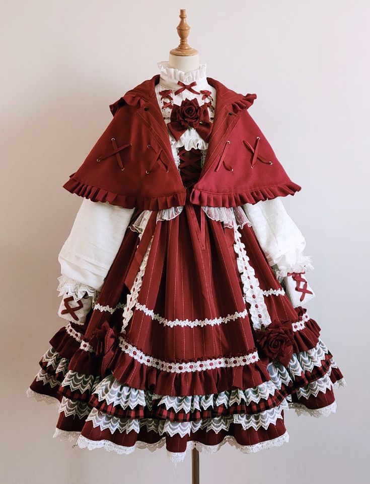 a fulllength image of a red and cream lolita fashion dress displayed on a mannequin. the dress has a full skirt with multiple layers of lace and ruffles, and a matching cape with a hood.  the bodice is fitted and adorned with bows. the overall aesthetic is elegant and vintageinspired.