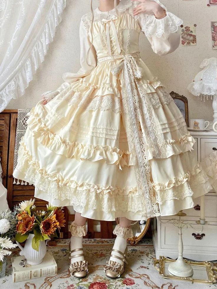 a woman is pictured in a brightly lit bedroom, wearing an elegant, tiered, offwhite lolita fashion dress with ruffles and lace accents. she is also wearing white lace arm warmers and white and gold doll shoes.