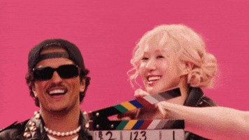 a gif image shows chaeyoung from the kpop group twice smiling happily while holding a film clapperboard with a man wearing sunglasses and a cap. they are in front of a pink background. the scene appears to be a behindthescenes shot from a film or music video production.
