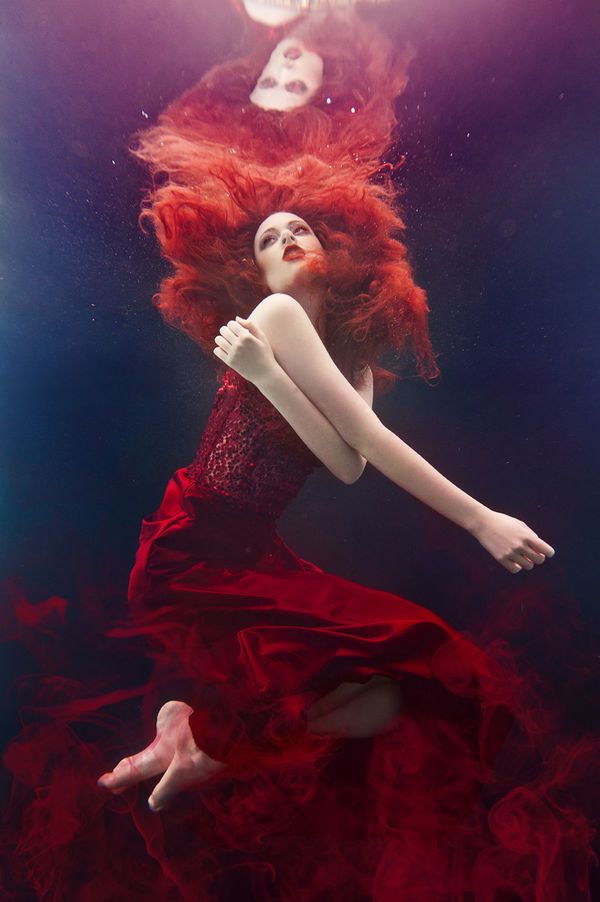 a woman with long red hair is floating underwater in a flowing red dress. her eyes are closed and she appears to be in a state of peaceful contemplation. the light coming from above casts a soft glow on her body and the flowing fabric of her dress.