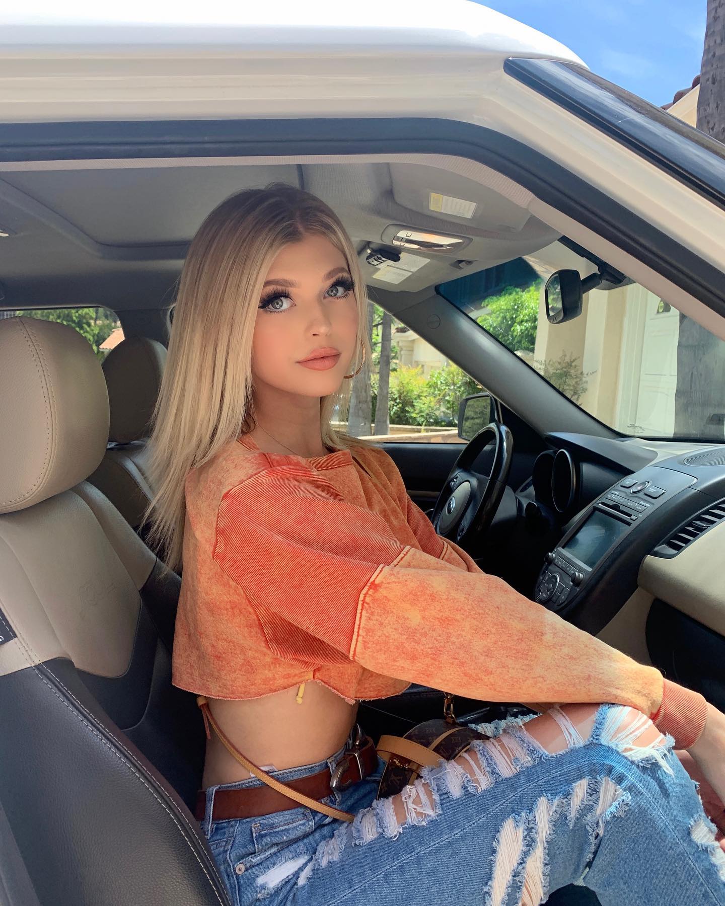 a blonde woman is sitting in a car. she has long blonde hair, wears an orange longsleeved top and ripped jeans. she also wears hoop earrings and a leather belt. a louis vuitton bag is visible in the image as well.