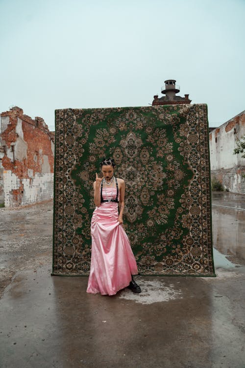 a young woman in a long pink satin dress stands in front of a large, ornate green carpet. the carpet is leaning against a dilapidated brick building. the woman is wearing black boots and jewelry. she has a slightly rebellious pose. the setting is urban and somewhat grunge. the ground is wet, indicating it might be raining. the overall mood is moody and stylish.