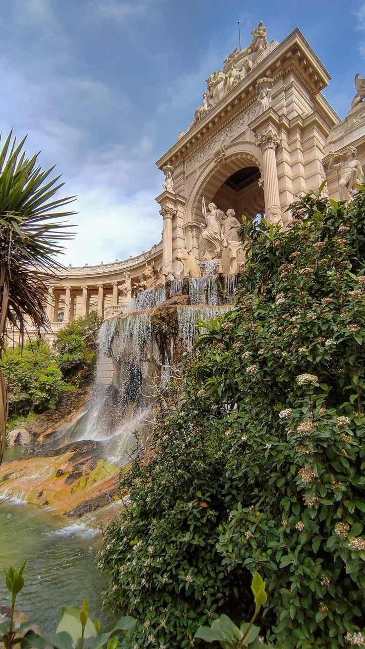 a captivating image of the cascading fountain at the palais longchamp in marseille, france.  the photograph showcases the grandeur of the palace's architecture, with its classical columns, intricate sculptures, and a large archway framing the waterfall. lush greenery and meticulously manicured bushes surround the fountain, enhancing the overall beauty and serene atmosphere of the scene. the water flows down the rocky terrain, creating a visually stunning effect. the sky is partially cloudy, adding depth to the background. the image highlights a harmonious blend of nature and human artistry.