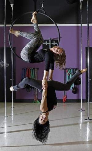 two women are performing an aerial silks routine. the woman on top is holding the hoop with her hands, while the woman on the bottom is holding onto the top woman's legs. they are both smiling and seem to be enjoying themselves. the background is a purple wall with a black ceiling.