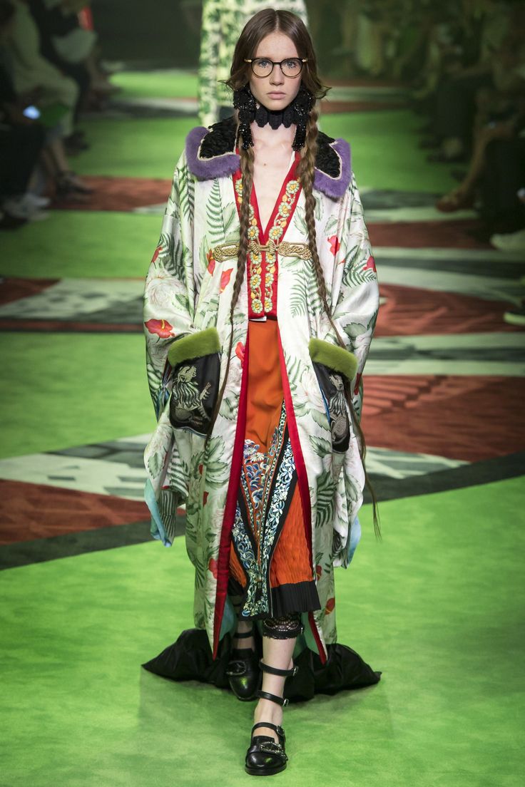 a model walks down a green runway during a gucci fashion show. she is wearing a long, floralprinted kimono with a fur collar, an orange skirt, and black shoes. her hair is in long braids, and she is wearing glasses. the overall style is eclectic and mixes different cultural and historical references. the background shows other people and a runway floor.
