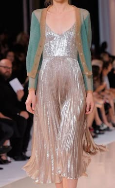 a woman in a silver pleated dress and a turquoise cardigan is posing for the camera on the runway. she's surrounded by other people, and the overall scene is elegant and glamorous.