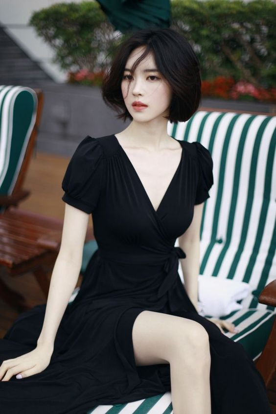 a young woman with short black hair is sitting on a striped deckchair. she is wearing a black vneck dress. the image is taken outdoors, with natural lighting. the perspective is at eye level.