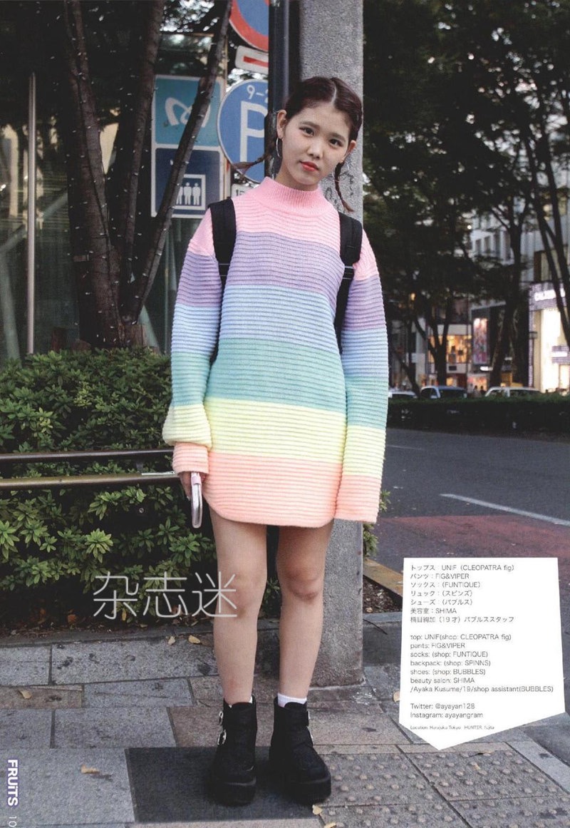 a young woman poses on a city sidewalk wearing a brightly colored, oversized rainbow sweater dress. she pairs the statement piece with black boots, a black backpack, and her hair pulled back in a simple style. the image captures a sense of effortless cool, reflecting a popular street style aesthetic.  the background suggests a bustling urban environment with trees and buildings partially visible. the overall tone of the image is bright and cheerful, due in part to the overcast lighting which provides a soft, even illumination.