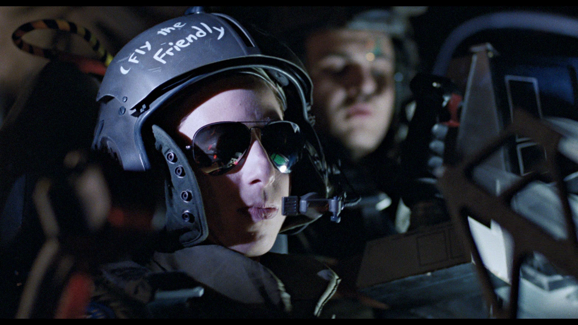 a pilot wearing a black helmet with "fly the friendly" written on it and sunglasses, sits in the cockpit of a helicopter, looking ahead at the controls. there is a blurred figure in the background in another seat. the image is dark and moody, and the pilot's expression is serious and focused.