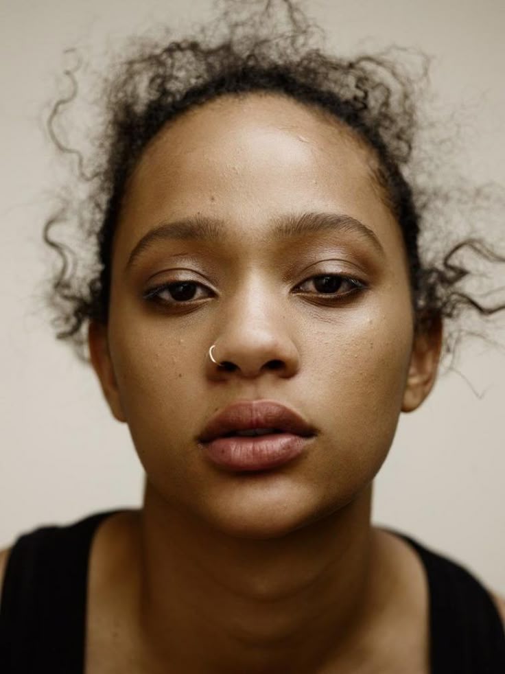 a closeup portrait of a woman with curly hair and a nose ring. she has dark eyes and a serious expression, looking directly at the viewer. the lighting is natural and soft, emphasizing her features. she is wearing a black tank top and minimal makeup. the background is plain and neutral, focusing attention on her face.