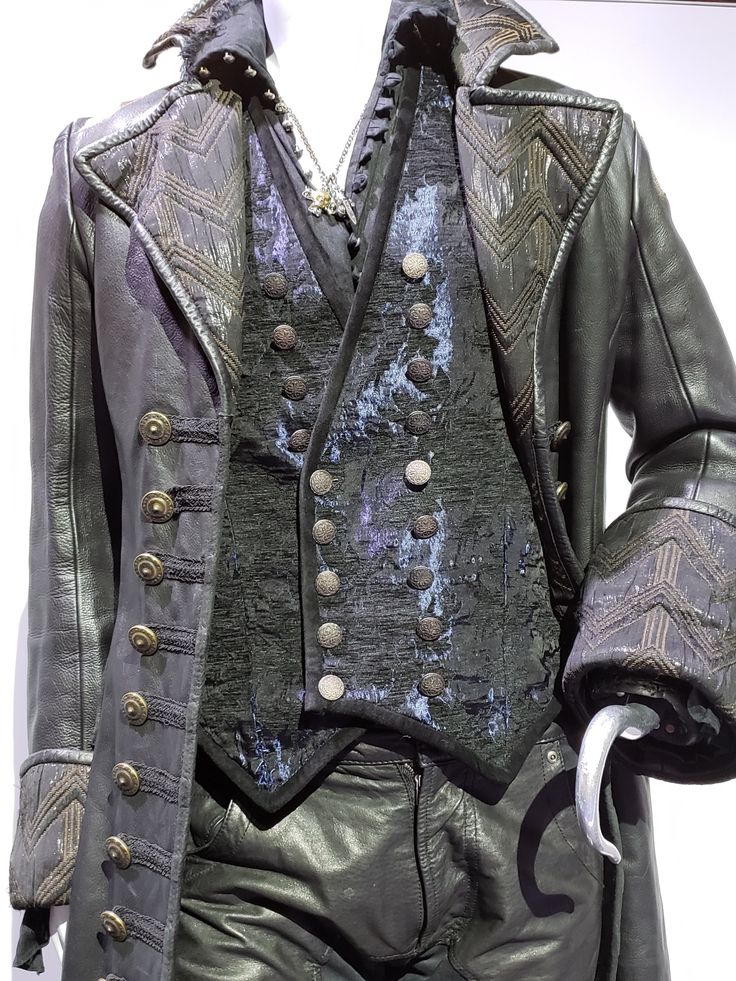 the photo shows a closeup of captain hook's costume from the tv show once upon a time. the costume consists of a long black leather coat with decorative trim, a dark vest with silver buttons, and shiny black leather pants. hook's signature hook is visible on the right sleeve of the coat. the outfit appears to be displayed on a mannequin.