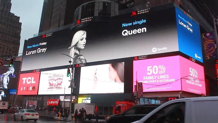 the image shows a vibrant and bustling scene of times square in new york city, dominated by large electronic billboards displaying various advertisements. prominently featured is a billboard advertising loren gray's new single "queen" on spotify, alongside the marriott marquis logo. other advertisements include levi strauss & co., tcl, and a 50% off promotion for family lines for military and veterans. the image captures the dynamic energy of times square, with its bright lights, numerous pedestrians, and passing vehicles.