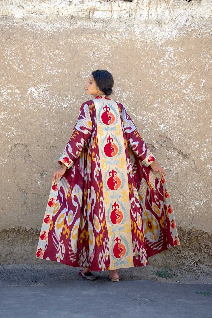 a back view of a woman wearing a long, flowing uzbek ikat robe. the robe is predominantly red with beige and gold ikat patterns and features a central panel with embroidered pomegranate motifs. the woman is standing outdoors with a textured, earthytoned wall in the background.  the image showcases the intricate details of the handcrafted garment and its rich cultural heritage. the overall aesthetic is elegant and stylish.