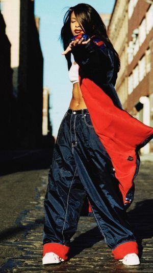 a full shot of aaliyah, a renowned 90s hiphop artist, standing on a cobblestone street.  she's dressed in iconic 90s streetwear: baggy dark blue nylon pants with red cuffs, a red and black jacket worn open, and white shoes.  her long dark hair is partially covering her face. she strikes a pose,  her hand gesturing towards the camera. the background is a city street with buildings visible. the image evokes the style and atmosphere of the 1990s.