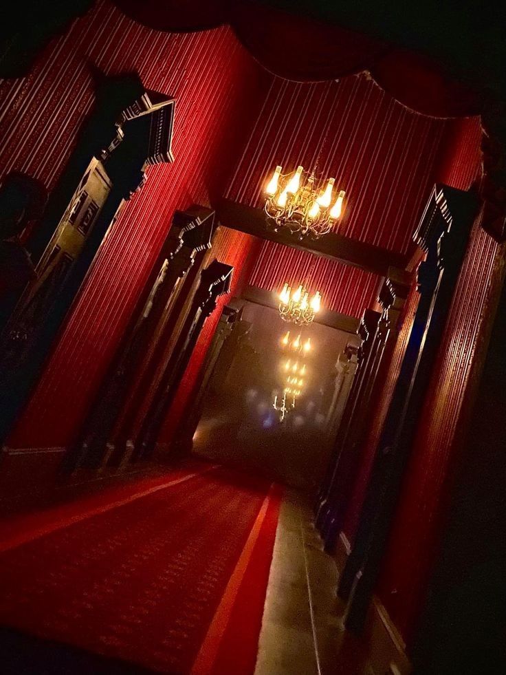 a dimly lit, dark hallway with a red carpet stretching down its length. the walls are lined with dark wooden panels and the ceiling is adorned with a patterned wallpaper. two ornate chandeliers hang from the ceiling, casting a warm glow on the red carpet. the atmosphere is eerie and mysterious, with shadows dancing on the walls. the hallway seems to disappear into the darkness at the end. the image evokes a sense of grandeur and mystery.