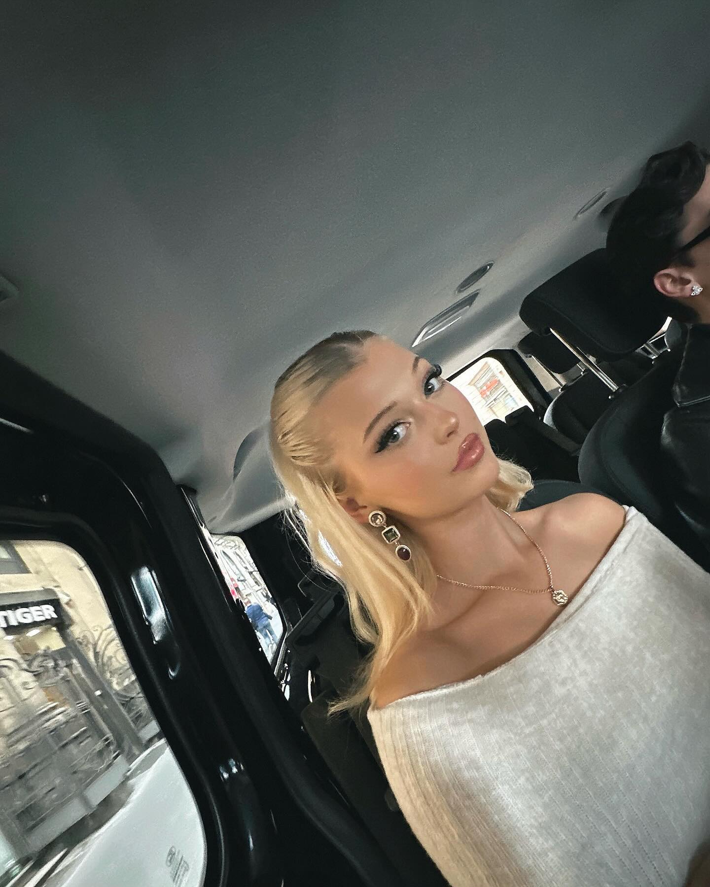 a woman with blonde hair is sitting in the passenger seat of a car. she is wearing earrings and a necklace, and an offtheshoulder top. the black car door and window are visible, with a city street and reflection seen through the window. in the background, a man is also sitting in the car. the woman has glossy lips.