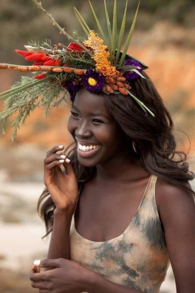 a closeup portrait of a beautiful black woman with a floral crown on her head, smiling happily. the woman's dark skin is glowing, and she has long, flowing hair. she is wearing a lightcolored tank top, and the background is blurred, focusing attention on her radiant smile. the image celebrates beauty, diversity, and natural radiance.