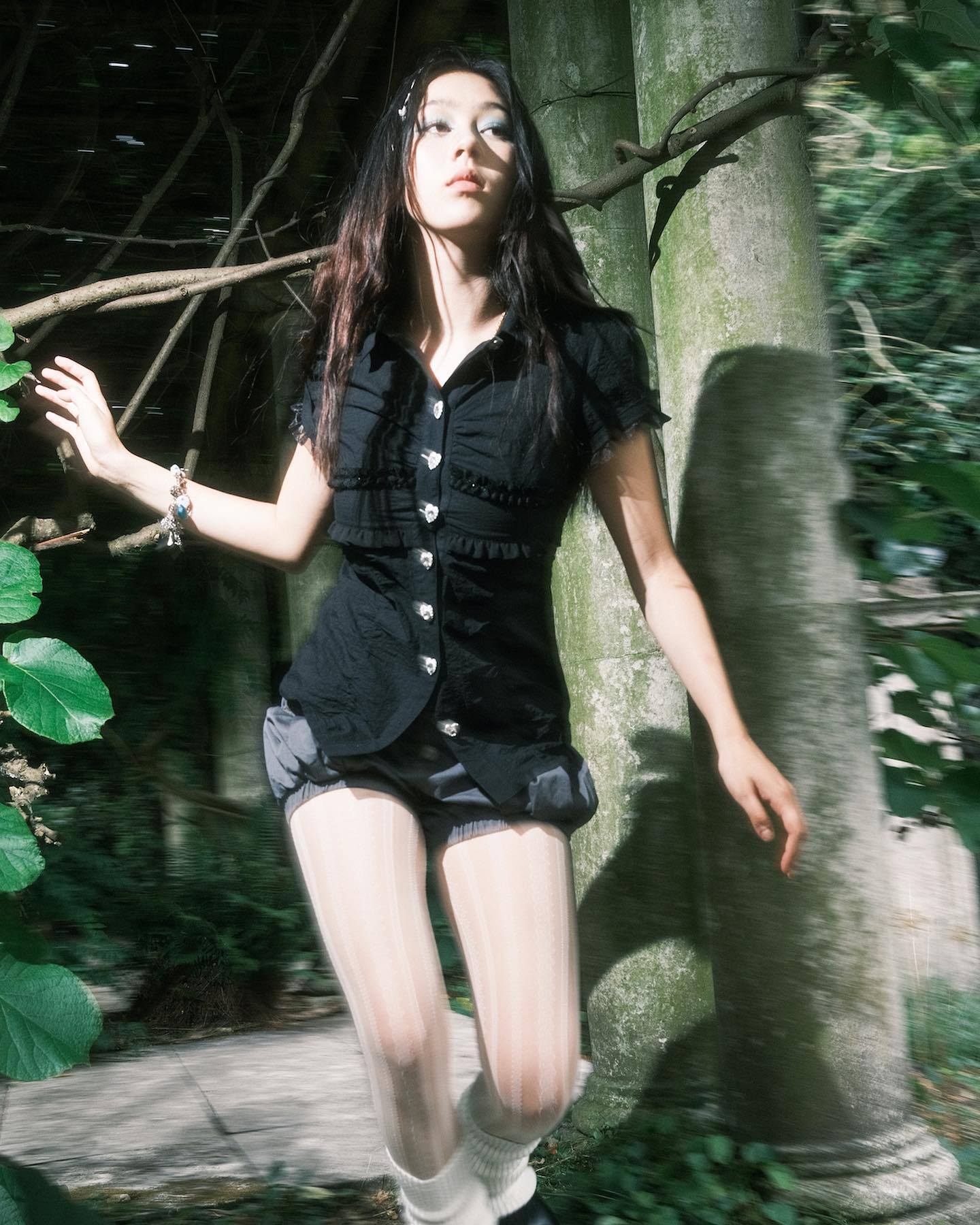 a young woman, possibly a model, is captured in a dynamic, motionblurred shot.  she's wearing a black buttondown shirt with puffed sleeves and short, dark grey shorts.  she also sports striped white tights, white ankle socks, and dark shoes (partially visible).  her long dark hair is loose, and she's wearing a bracelet.  the setting appears to be a garden or park, with lush greenery and aged, mossy pillars in the background. the overall aesthetic is a blend of dark and whimsical elements, with a hint of vintage or retro fashion.  the photo seems deliberately stylized, suggesting a fashion editorial or artistic concept.