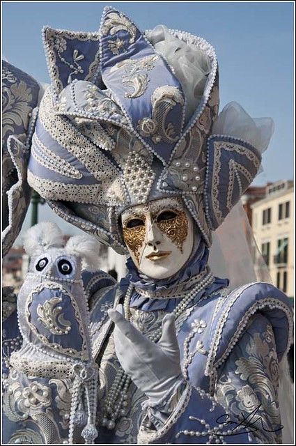a close up of a woman wearing an elaborate venetian masquerade costume. she has an ornate white mask with gold accents. the costume is blue and white with ornate details and pearls.  she is holding a white owl puppet.