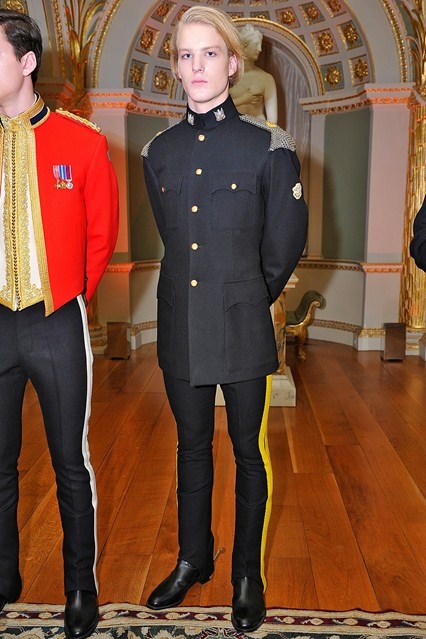 a man with blonde hair is standing indoors wearing a black military uniform with gold buttons and trousers with a yellow stripe down the side. he is wearing black dress shoes. another person is standing to his left wearing a red military uniform. the background includes ornate gold decorations and a wooden floor.