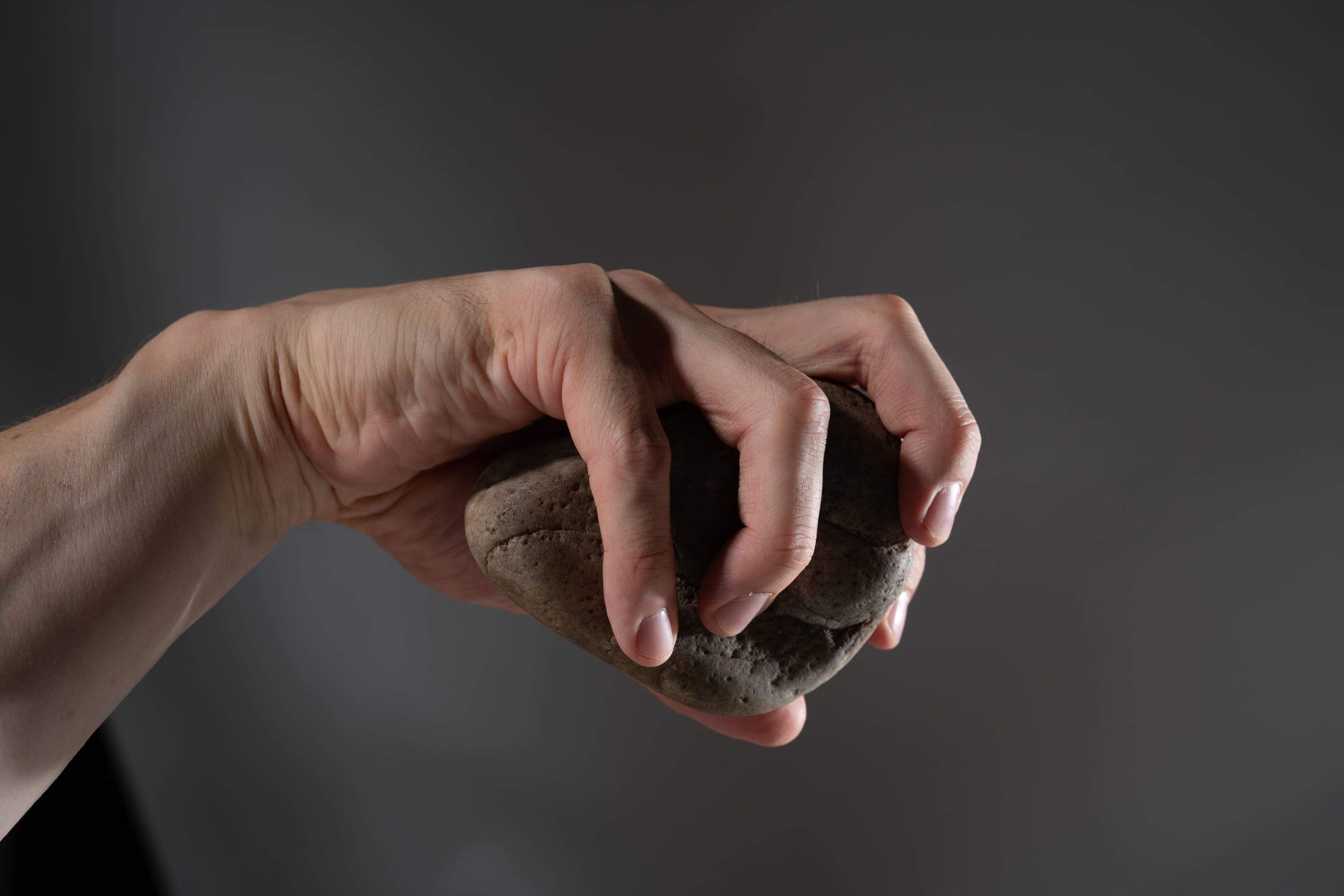 a hand is tightly gripping a brown rock. the hand is well lit against a dark background, with the focus on the texture of both the hand and the stone.