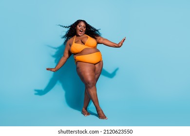 a black woman with curly hair is wearing a yellow bikini and smiling with her hands out on a blue background.  she is expressing happiness and joy in her own body.