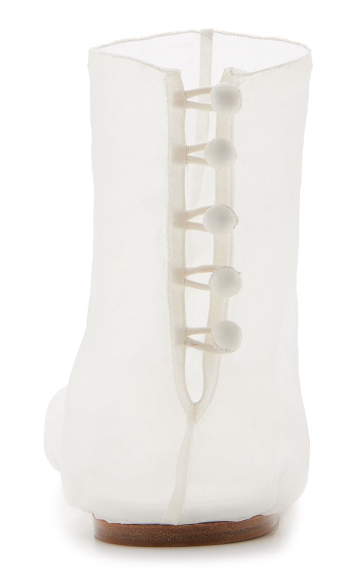 single, white sheer organza ballet flat is shown from a side profile view. the shoe features a unique back closure detail with small, lightcolored buttons along the back seam. the shoe has a slightly rounded toe and a simple, minimalist design. the sole appears to be made of wood. the overall aesthetic of the shoe is delicate, elegant, and light.
