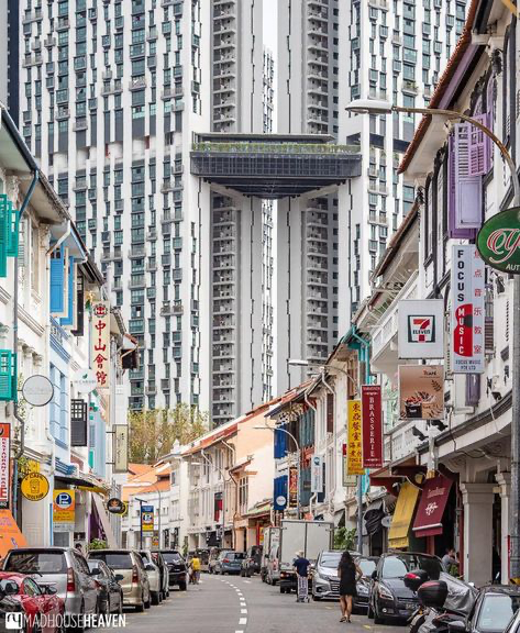 the image showcases a striking contrast between modern highrise architecture and traditional shophouses in singapore.  two towering, sleek, modern apartment buildings, connected by a skybridge, dominate the background.  these are juxtaposed with a narrow street lined with traditional twostory shophouses, displaying a mix of signage and businesses. the scene is a vivid example of the blend of old and new architecture often found in asian cities. the street is relatively quiet, with some pedestrians and parked cars adding to the scene's everyday feel. the image is wellcomposed, emphasizing the architectural contrast and the depth of the street perspective.