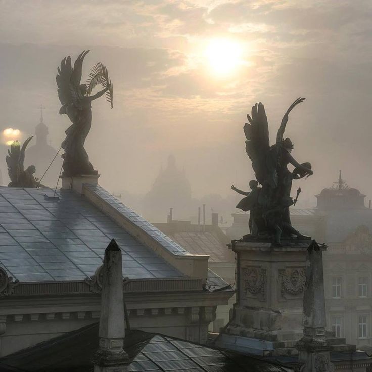 the image showcases a stunning cityscape shrouded in a thick, early morning fog.  the sun is rising, casting a soft, hazy light on the scene.  dominating the foreground are several large, ornate angel statues perched atop the roofs of buildings, their details slightly obscured by the mist.  the architecture appears to be of a european style, possibly baroque, with intricate rooflines and decorative elements visible through the fog.  the overall atmosphere is serene, mystical, and evocative, suggesting a moment of quiet beauty and mystery within a historic city. the fog adds a significant layer of depth and visual interest, emphasizing the statues and drawing the eye towards the distant, indistinct skyline.