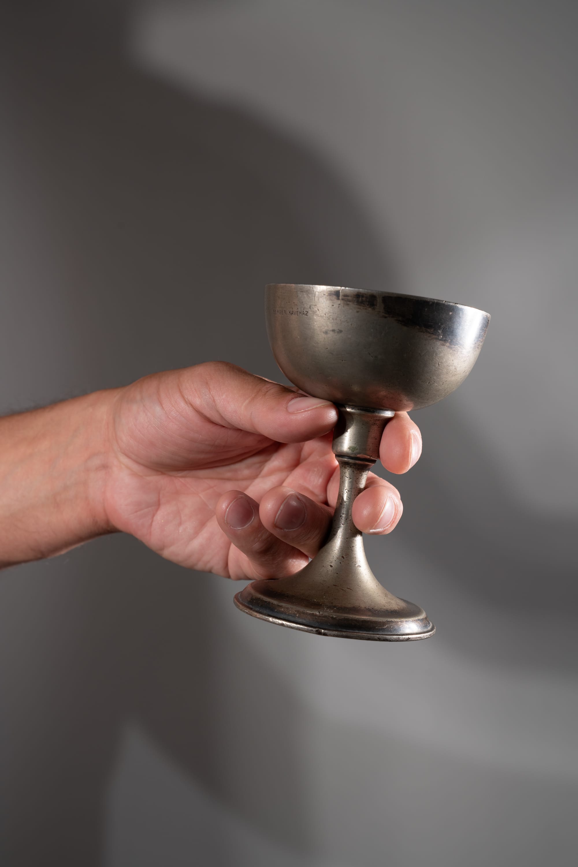 an adult hand holds a shallow, stemmed silver cup against a plain gray background. the cup is tarnished and has visible markings indicating its age and possible use. the studio lighting creates a strong contrast between the object and the background, emphasizing the cup's details and texture.