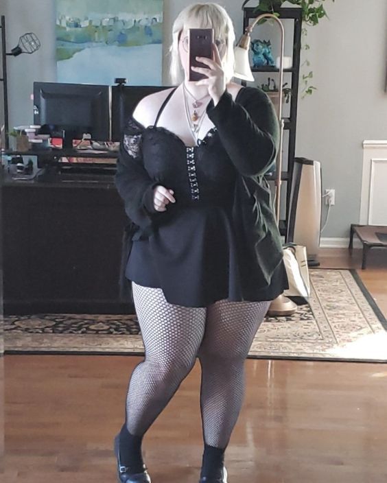 a woman in a black dress, fishnet tights, and black shoes stands in a home and takes a selfie. she is wearing a black lace top and a black cardigan with a necklace and rings. her phone is in her right hand, and she is looking at the camera. she has her hair pulled back, and she is wearing black eyeshadow and eyeliner.  the floor is wood, and the home is stylish and modern.