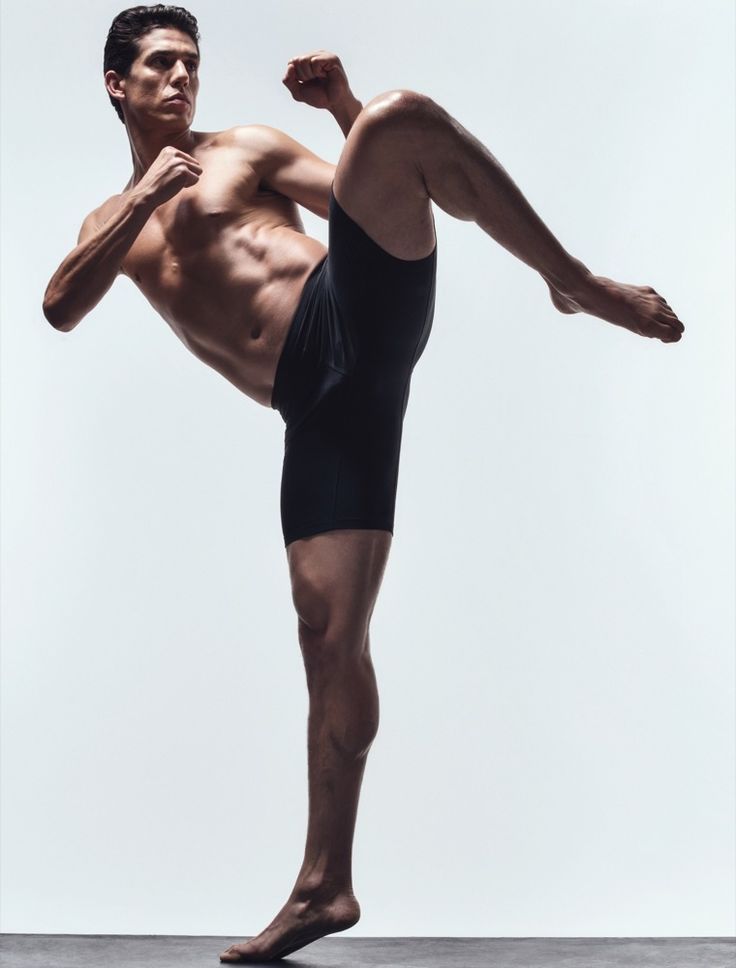 a young muscular man in black shorts is seen in a fighting stance. his right leg is lifted high in the air with his left leg planted firmly on the ground, as he prepares for a kick. his face is focused and determined. this image could represent power, strength, and the pursuit of fitness.