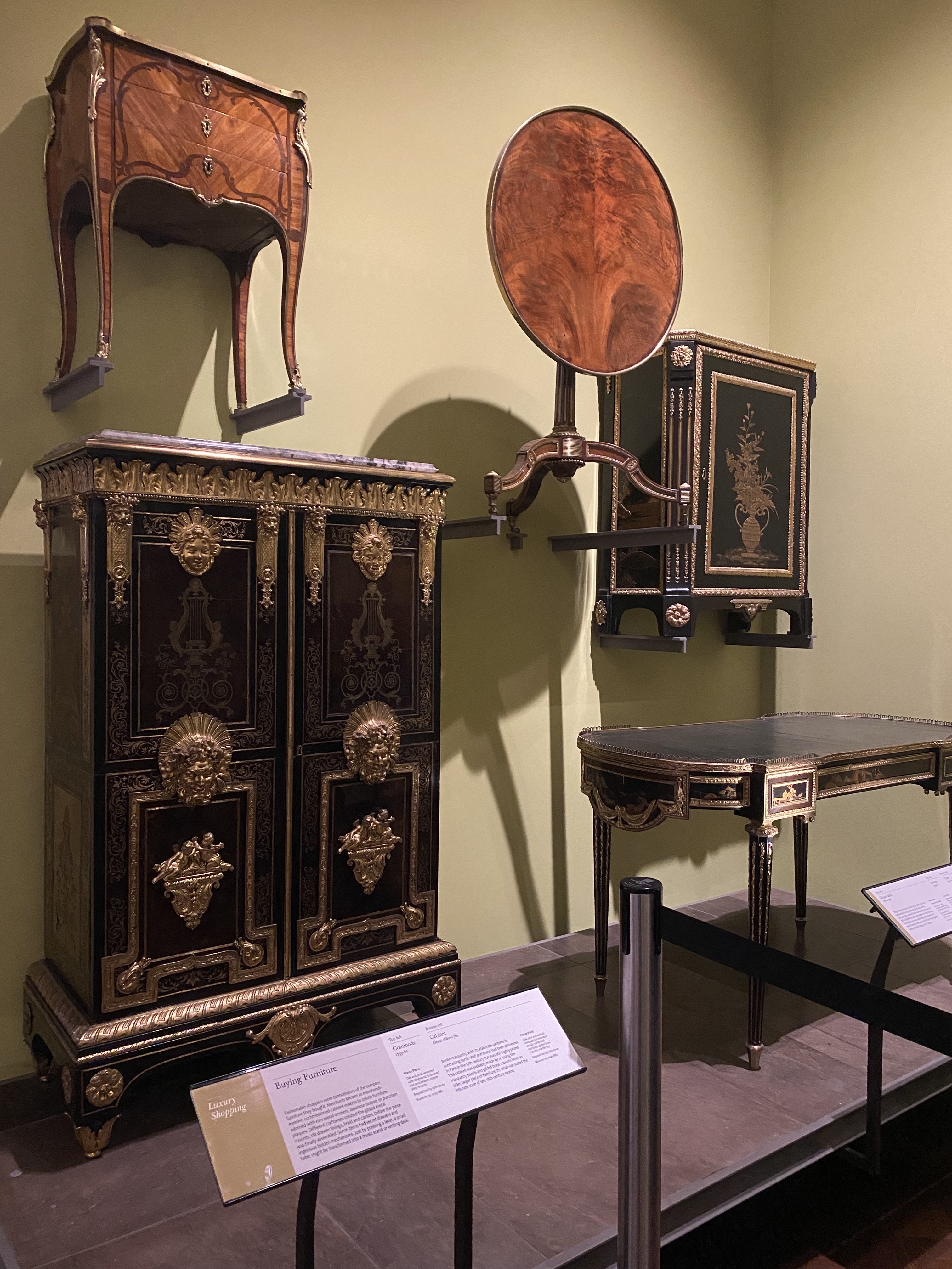 the image shows a collection of antique luxury furniture on display in a museum exhibit. there is a large black cabinet with gold trim and decorative elements, a small brown drawer cabinet mounted on the wall, a round wooden table attached to a black cabinet with gold trim, and a small black desk with gold trim. signage is present in front of the furniture. the furniture is displayed against a light green wall. a black rope barrier is visible in the foreground.