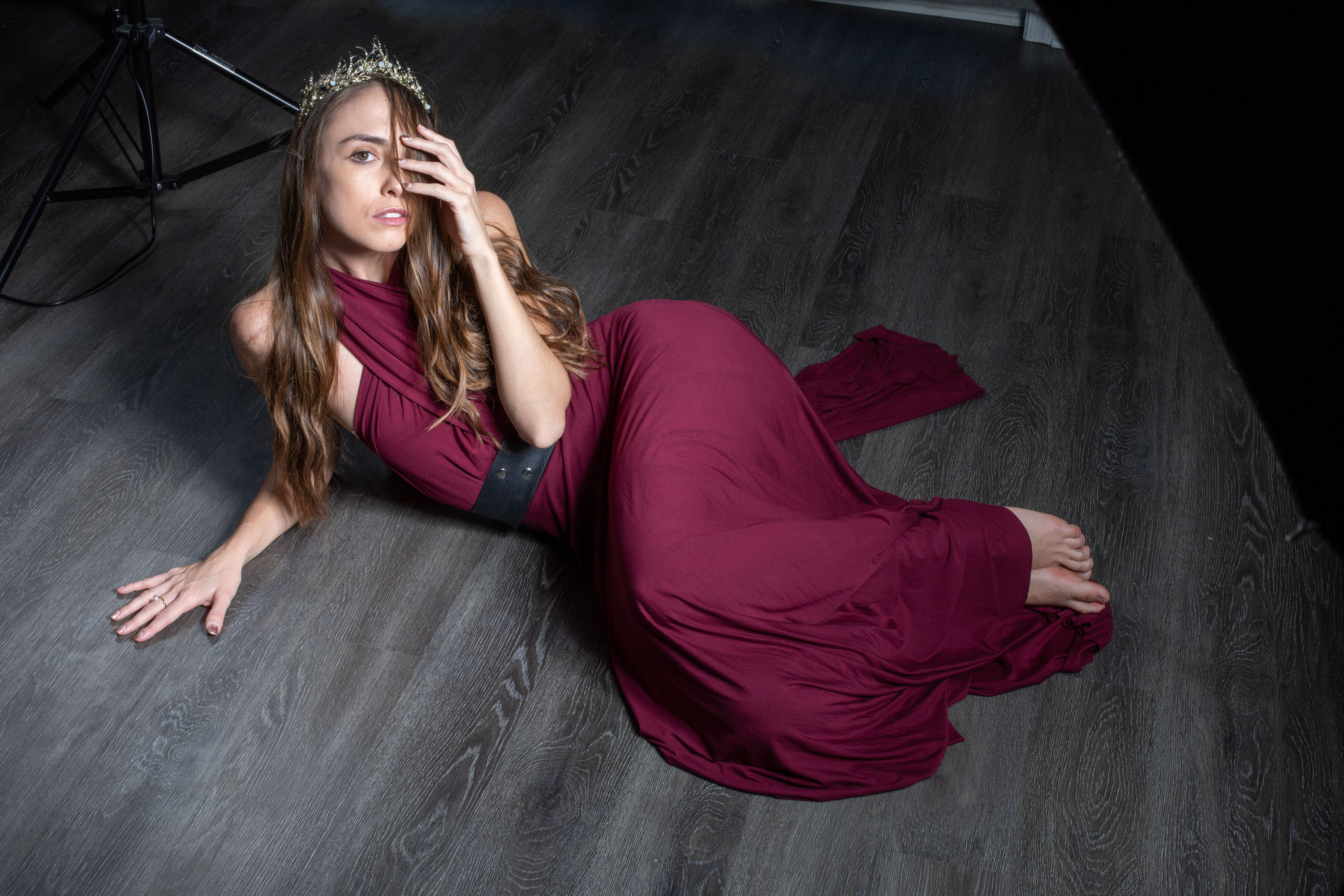 a young woman in a long maroon dress and a crown lays on a dark wooden floor in a studio. she is partially illuminated by studio lighting as she poses for a photoshoot.