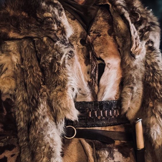 a closeup shot shows a person wearing a fur garment, likely a cloak or coat. the garment is adorned with various furs and pelts, giving it a medieval or fantasy appearance. the person is wearing a leather belt with a ring and knife. the image suggests a character in a historical or fantasy setting, perhaps a warrior or hunter.