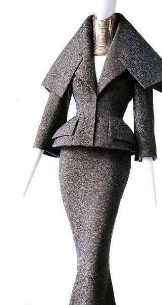 a mannequin is wearing a gray tweed suit, consisting of a peplum jacket with a wide collar and a fitted midi skirt. the mannequin is also wearing a large neckpiece.