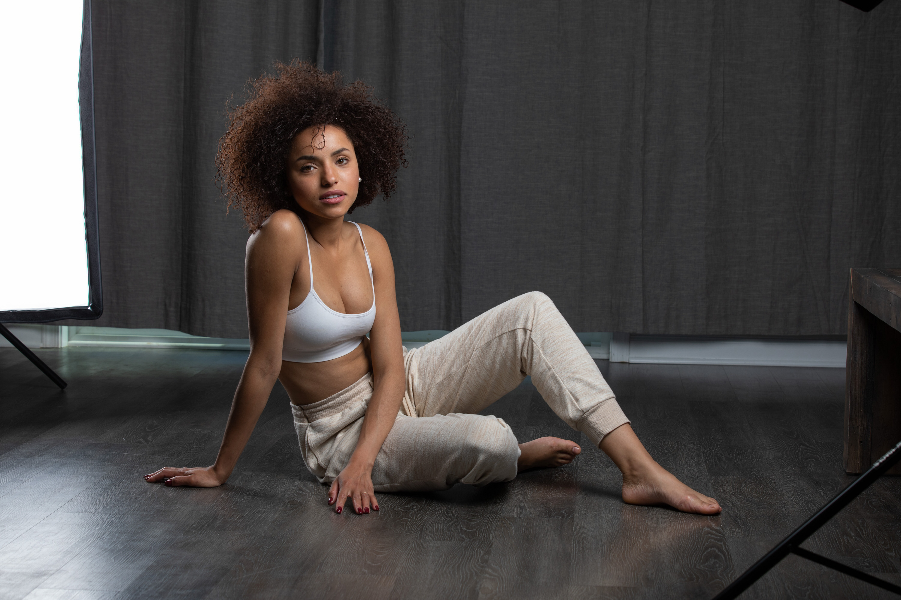 a woman with curly hair is sitting on the floor in a studio setting, wearing a white crop top and light brown sweatpants. she is looking directly at the camera with a relaxed and confident expression. the image is welllit, with soft light coming from a softbox on the left and natural light coming from a window offcamera. the background is a neutral gray color, and the overall mood of the image is casual and comfortable.