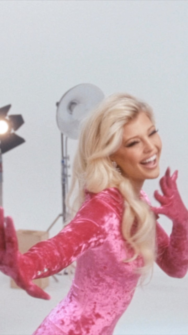 a woman with blonde hair, wearing a pink velvet dress and matching gloves, is smiling in a photography studio with studio lights in the background.