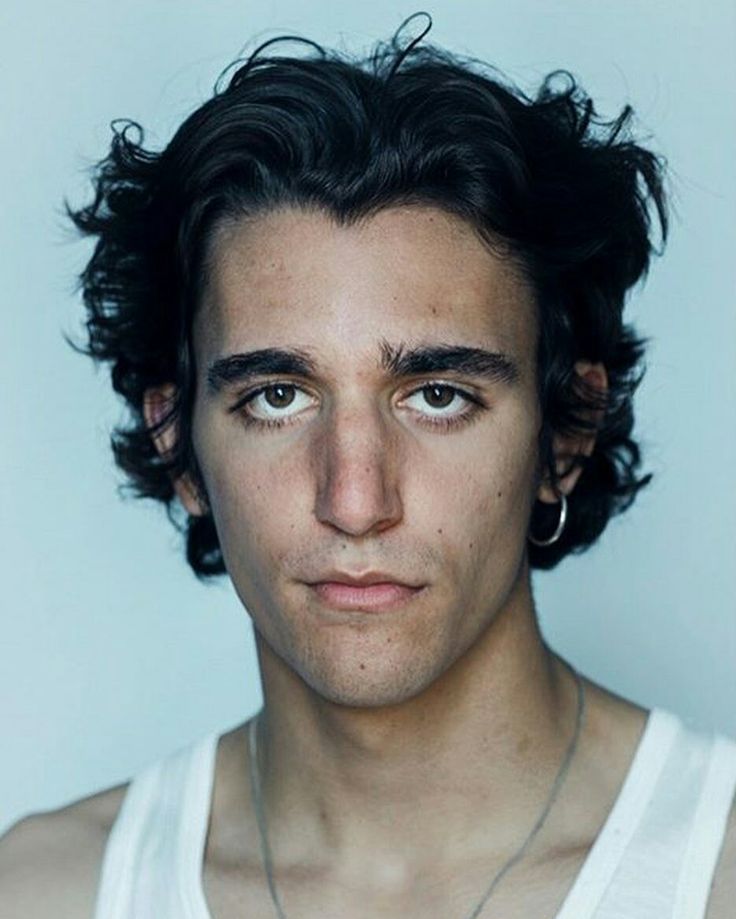 a closeup portrait of a young man with dark, curly hair. he has olive skin and a serious expression. he is wearing a white tank top and a simple necklace. the background is a neutral, light blue. the image is welllit and appears to be a professional studio shot. the man looks directly at the camera.
