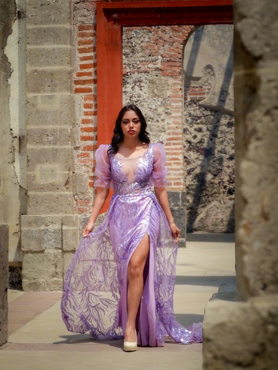 a woman in a long purple dress with a high slit stands in a doorway made of stone and brick. she poses confidently, holding the sides of her dress. the dress has shiny sequins and a delicate pattern. she is wearing high heels that match the dress. the building is old and the stone is weathered.