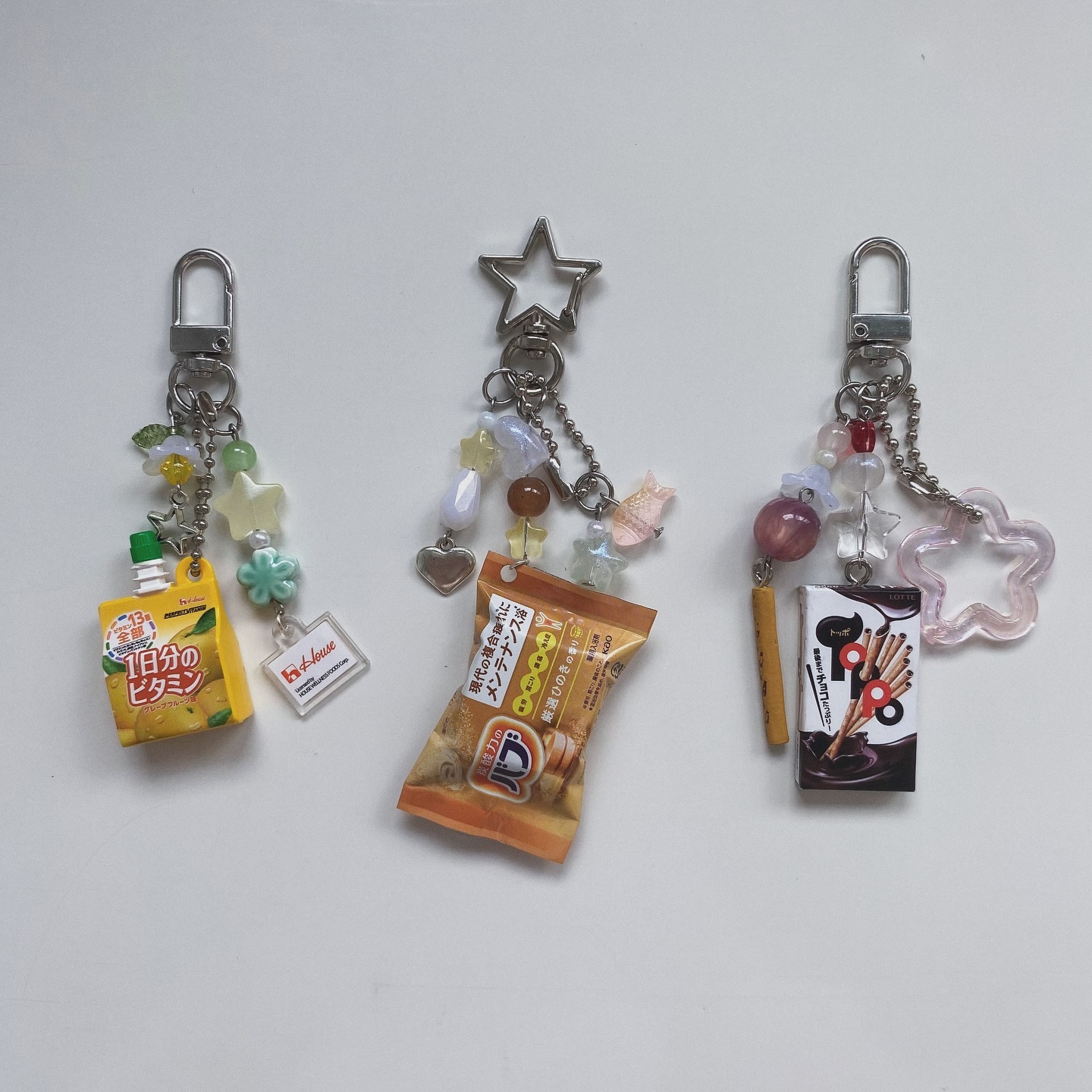 the image shows three keychains with miniature food charms and decorative beads. the keychain on the left features a miniature of a vitamin drink, the one in the center has a cracker snack charm, and the keychain on the right has a chocolate sticks charm. all three keychains have multiple decorative beads and metal clasps.